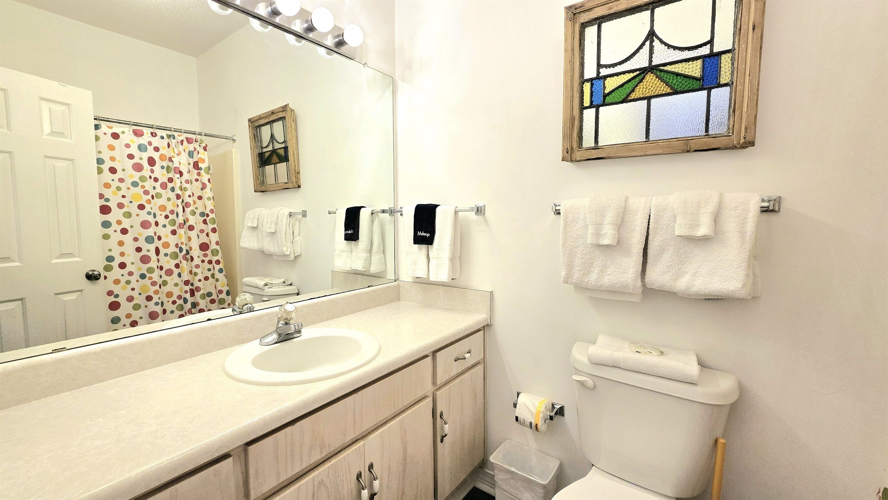 A bathroom with a sink, mirror, and toilet. There are towels hanging on the wall and a shower curtain with a polka dot pattern.