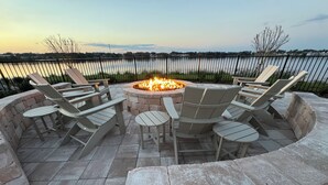 An outdoor seating area with Adirondack chairs around a stone fire pit, overlooking a body of water.