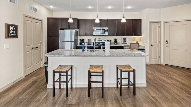 A modern kitchen with dark cabinets, a stainless steel refrigerator, a microwave, and a large island with bar stools.