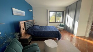 A modern bedroom with a bed, a green chair, a white ottoman, and a window with a view of greenery. - Romantic mini villa - heated pool and wooded park -Les Hauts de Locmiquel (Baden)