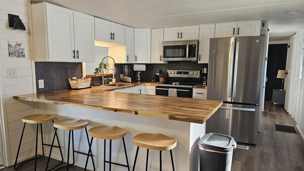 A modern kitchen with white cabinets, a wooden island with bar stools, stainless steel appliances, and a trash bin. - Slice of Paradise w/community pool near beaches & downtown! (Fort Myers)