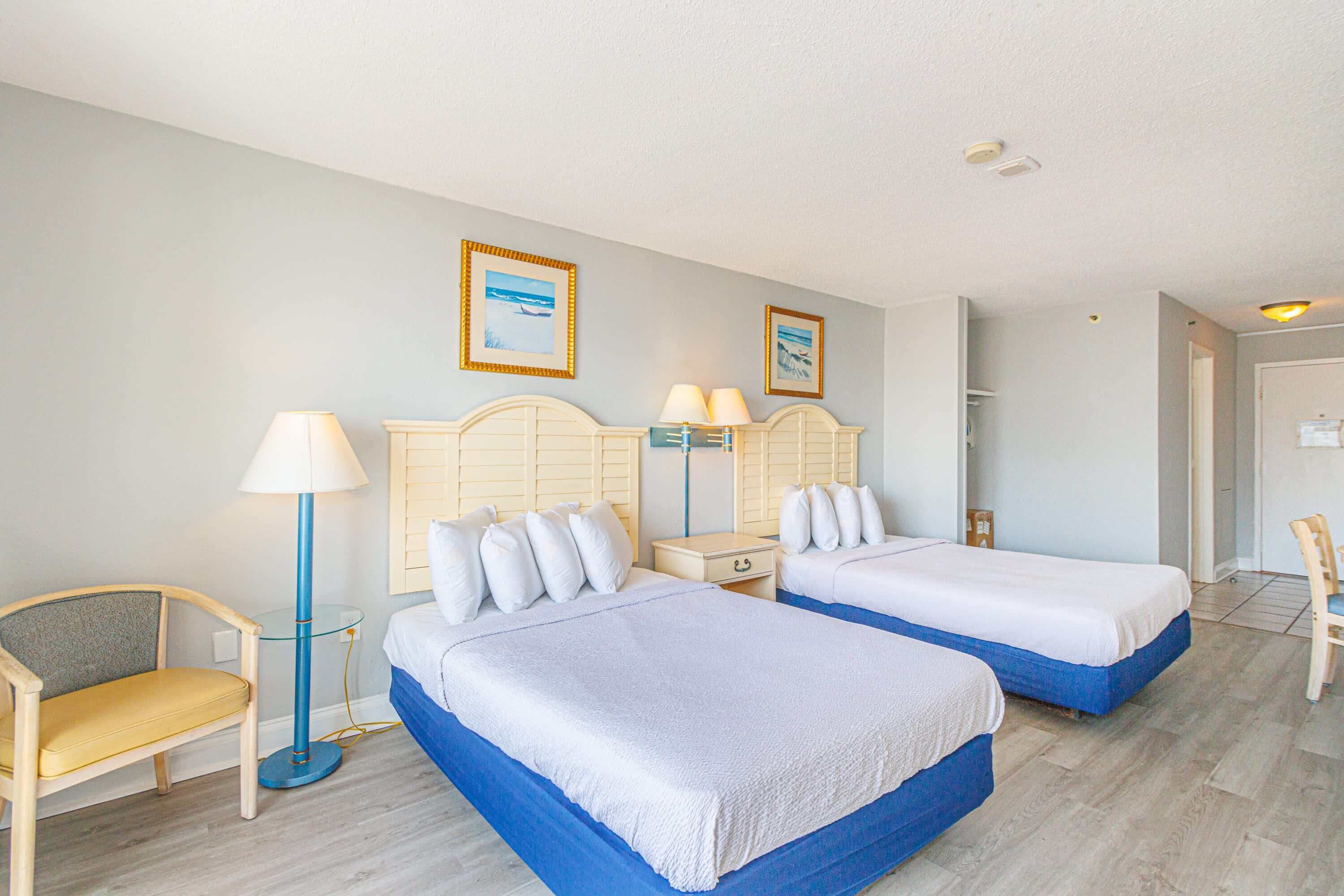 A hotel room with two beds, a chair, a lamp, and framed pictures on the wall.