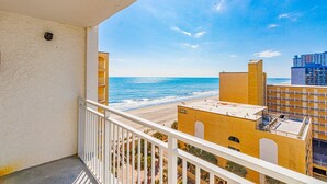 Ein Balkon mit Blick auf Strand und Meer, ein Gebäude mit gelber Fassade und ein klarer Himmel.