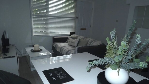 A modern living room with a white sofa, a small side table with a candle, a white coffee table with a black placemat, and a potted plant.
