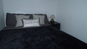 A neatly made bed with a black blanket, two pillows with a text design, and a bedside lamp on a nightstand.