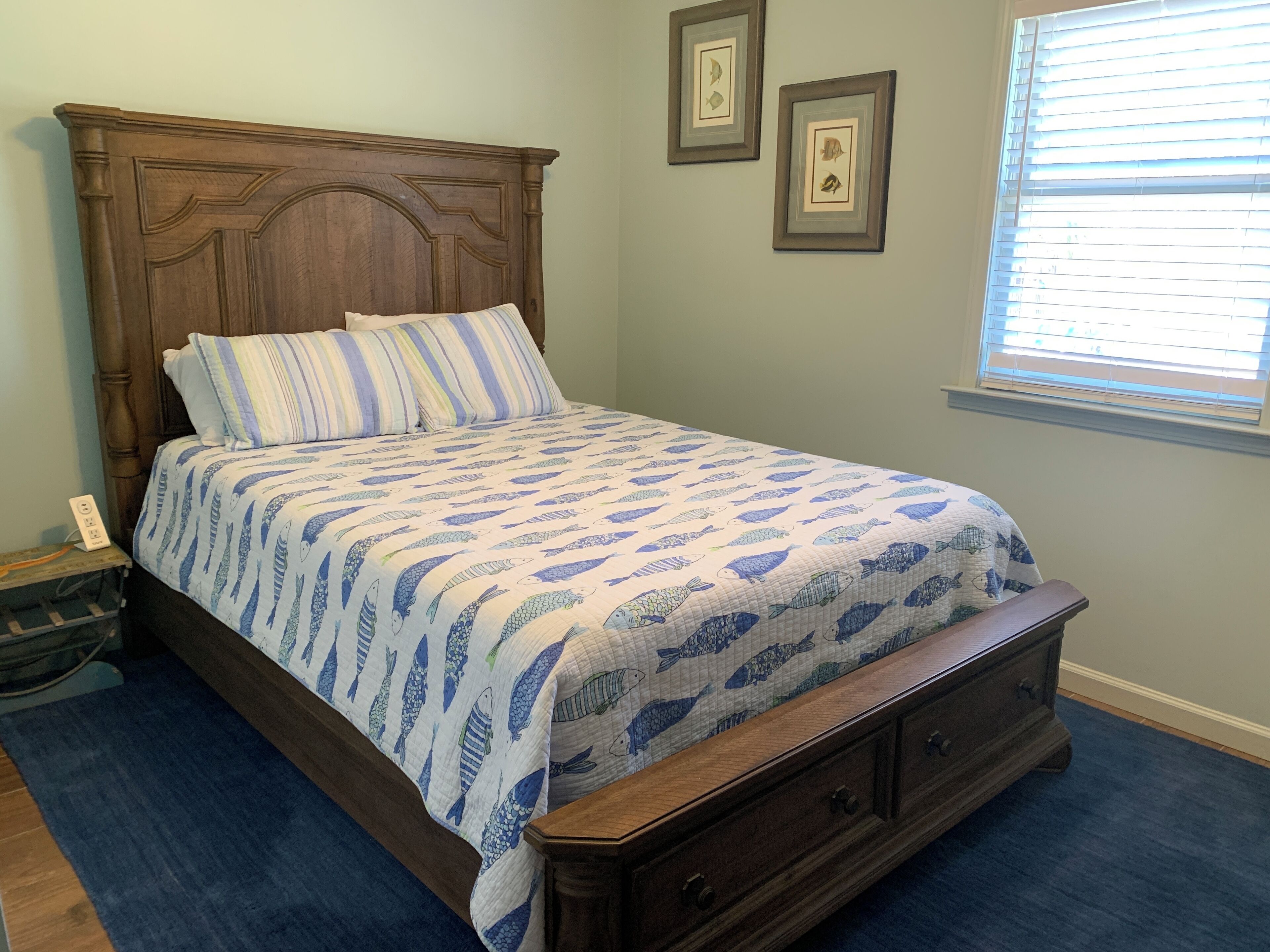 A bedroom with a wooden bed, striped pillows, a quilt with fish patterns, a nightstand, and a window with blinds.