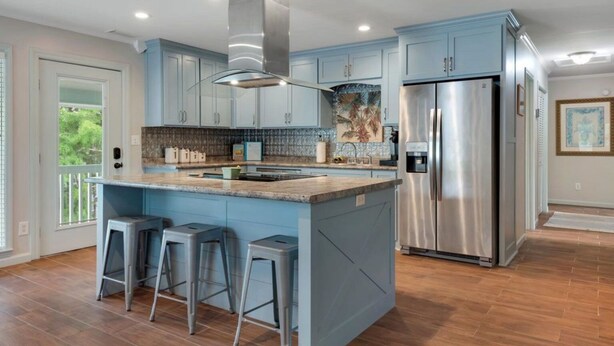 A modern kitchen with blue cabinets, a stainless steel refrigerator, and a central island with bar stools. - 3-bedroom Riverfront home with private boat dock and view of Steinhatchee River. (Steinhatchee)
