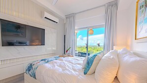 Une chambre dotée d’un grand lit, d’un téléviseur à écran plat fixé au mur, d’une fenêtre donnant sur un paysage verdoyant et d’un ventilateur de plafond.