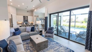 Ein modernes Wohnzimmer mit einer Couch, einem Couchtisch und Sesseln. Ein großes Fenster bietet Blick auf einen Pool und Gartenmöbel.