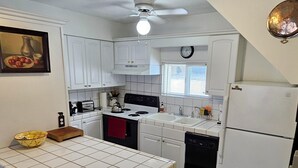 Une cuisine compacte avec des meubles blancs, une cuisinière, un évier et un réfrigérateur. Il y a des tabourets au comptoir et une fenêtre au-dessus de l’évier.