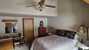Une chambre à coucher comprenant un lit, un bureau en bois, un miroir, un ventilateur de plafond et une table de chevet avec une lampe.