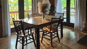 Een houten eetkamer met een grote tafel en stoelen, grote ramen met gordijnen en uitzicht op groen.