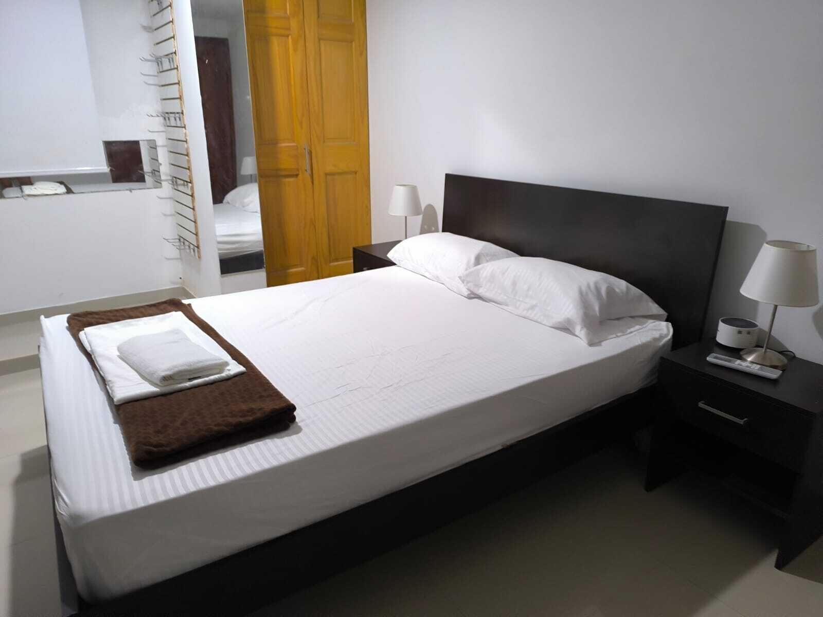 Una cama bien hecha con sábanas blancas y una manta marrón, una mesita de noche con una lámpara, y una puerta de madera al fondo.