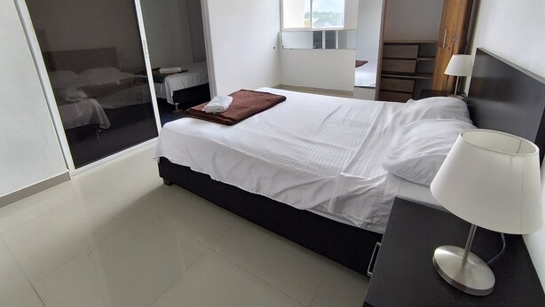 A modern bedroom with a large bed, a bedside table with a lamp, a wardrobe, and a sliding glass door leading to another room.
