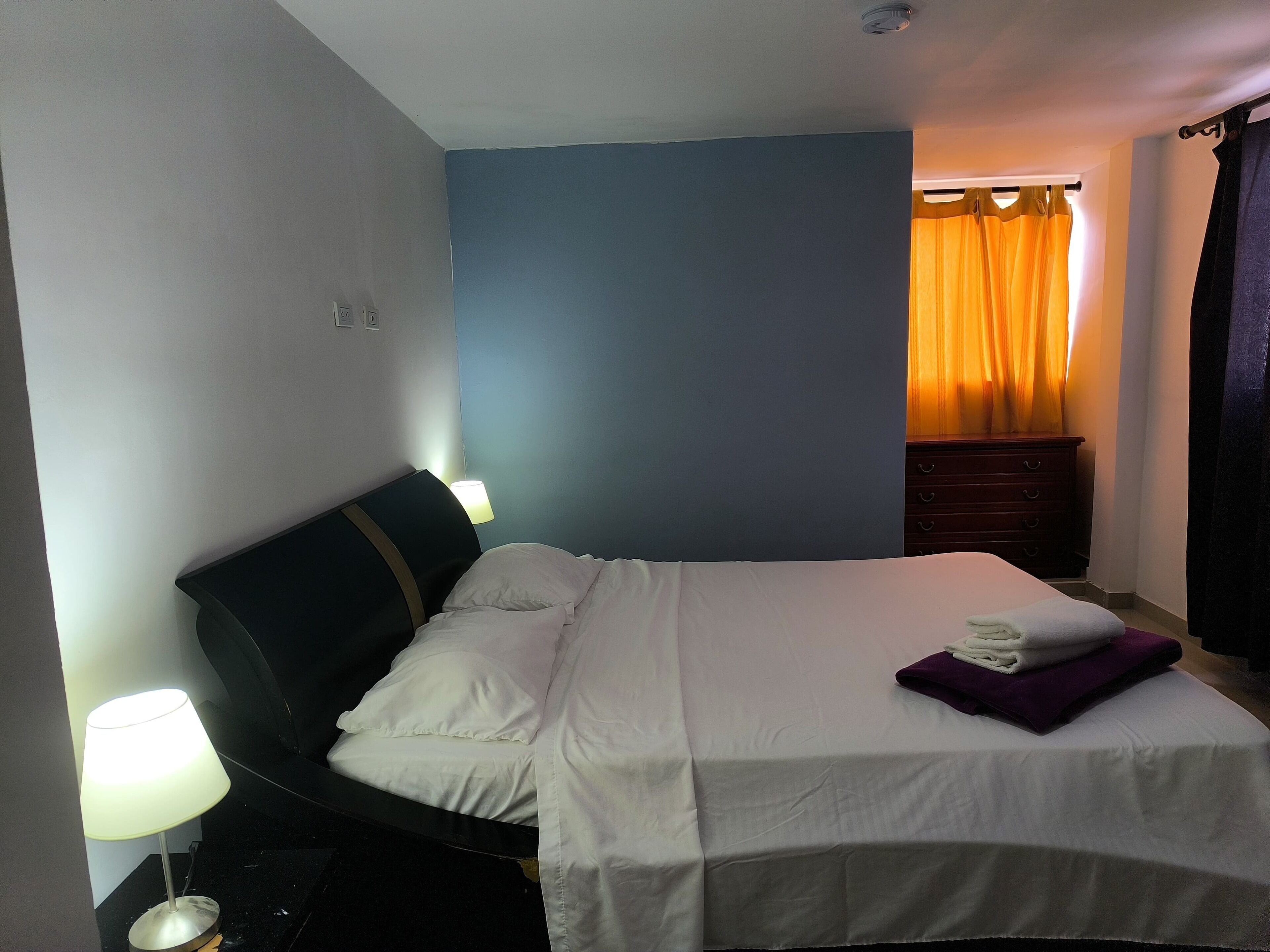 Una cama bien hecha con ropa blanca y una almohada morada, una lámpara de noche y un aparador de madera oscura frente a una pared azul.