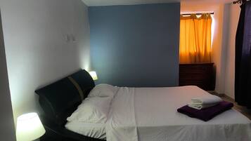 Una cama bien hecha con ropa blanca y una almohada morada, una lámpara de noche y un aparador de madera oscura frente a una pared azul.