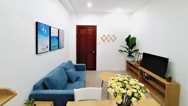 Een woonkamer met een blauwe bank, een salontafel, een televisie op een houten dressoir, en een deur op de achtergrond.