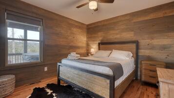 A bedroom with a wooden floor, a bed with a headboard, a nightstand, a bedside table, a window, and a ceiling fan.