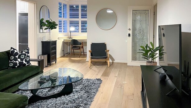 Ein modernes Wohnzimmer mit einem Glastisch, einem grünen Sofa, einem Holzstuhl und einem Fernseher auf einem schwarzen Standfuß.