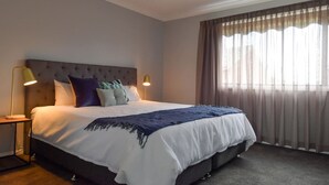 Een groot bed met een gestoffeerd hoofdeinde, twee bedlampjes en uitzicht op een stadsgezicht door dunne gordijnen.