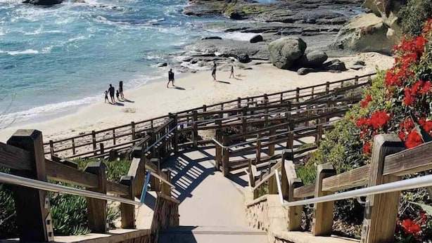 Un escalier en bois mène à une plage. Il est doté d’une rampe en bois et d’un mur de pierres sur un côté.