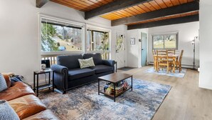 Amplia sala de estar con sofá de cuero negro, una mesa de centro y una mesa de comedor con sillas. Ventanas grandes permiten luz natural y vistas al exterior.