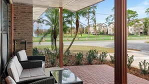 A covered patio with wicker furniture and a glass table, overlooking a residential street with houses and palm trees. - Stylish & Cozy King Bed Retreat @ Baldwin Park (Orlando)