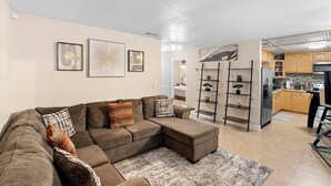 A modern living room with a sectional sofa, a coffee table, and a kitchen area with wooden cabinets and stainless steel appliances. - Stylish & Cozy King Bed Retreat @ Baldwin Park (Orlando)