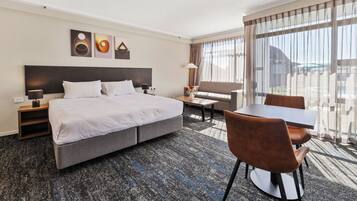 A hotel room with a large bed, a dining table, and a sofa. There are wall art pieces above the bed and a window with curtains.