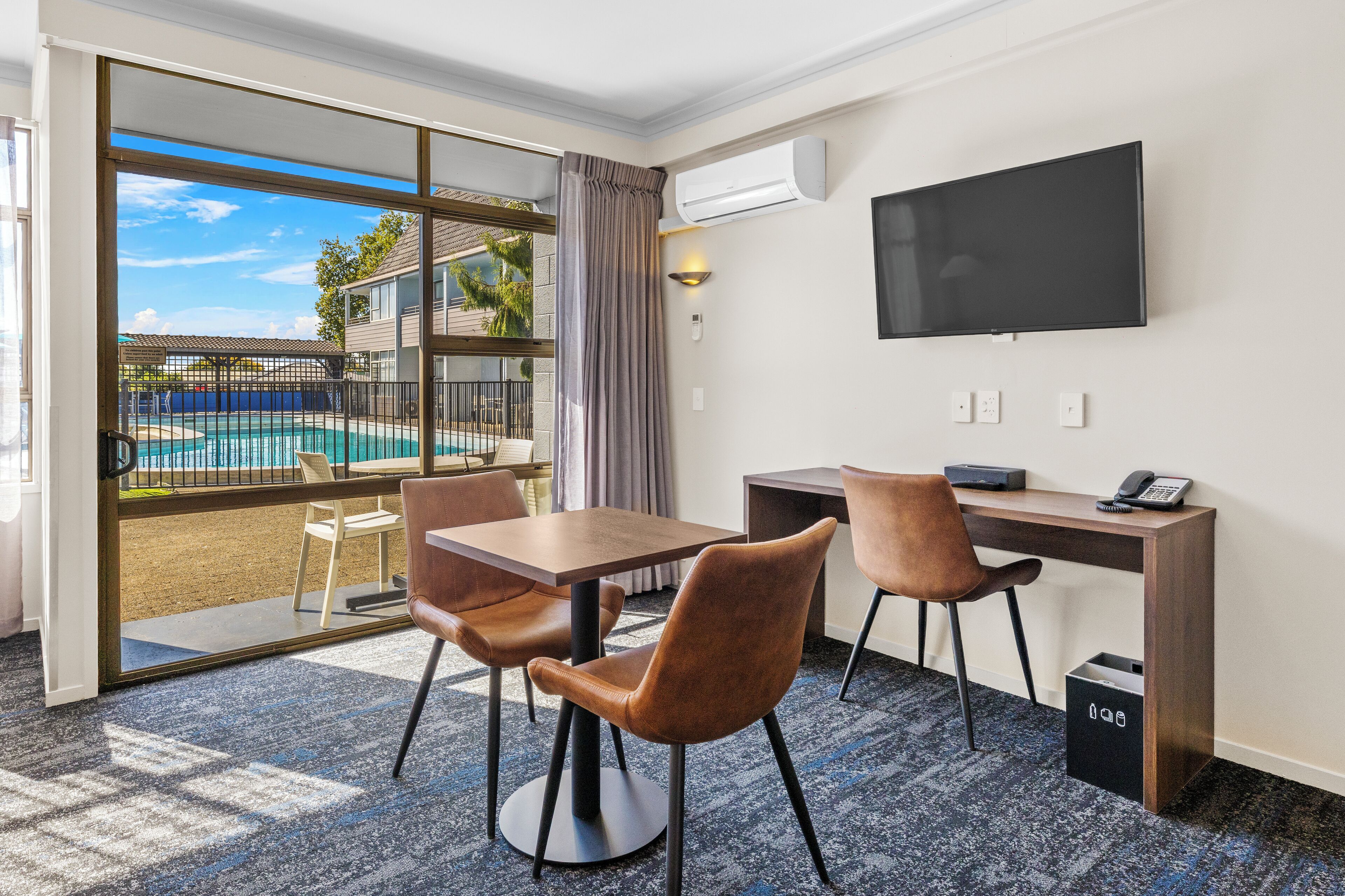A hotel room with a pool view, a desk with a chair, a TV, and an air conditioning unit.