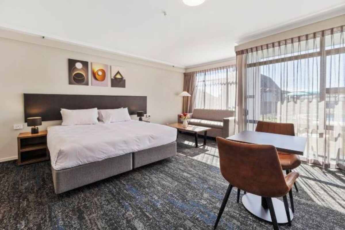 A hotel room with a large bed, a desk, and a chair. There are three paintings on the wall, and a window with curtains.