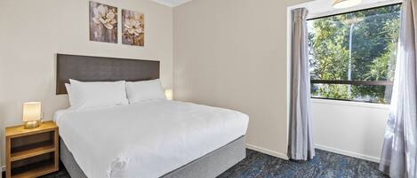 Habitación de hotel con cama, mesita de noche con lámpara, ventana con cortinas y una obra de arte en la pared.