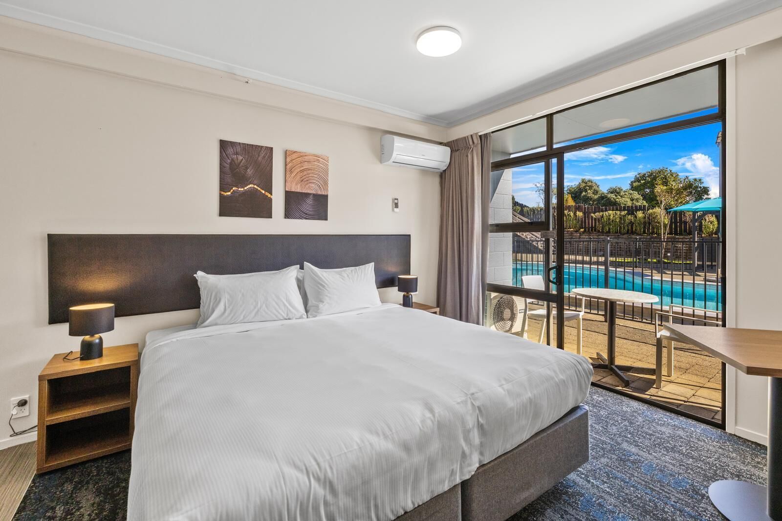 A hotel room with a large bed, a nightstand, a desk, and a view of a swimming pool.