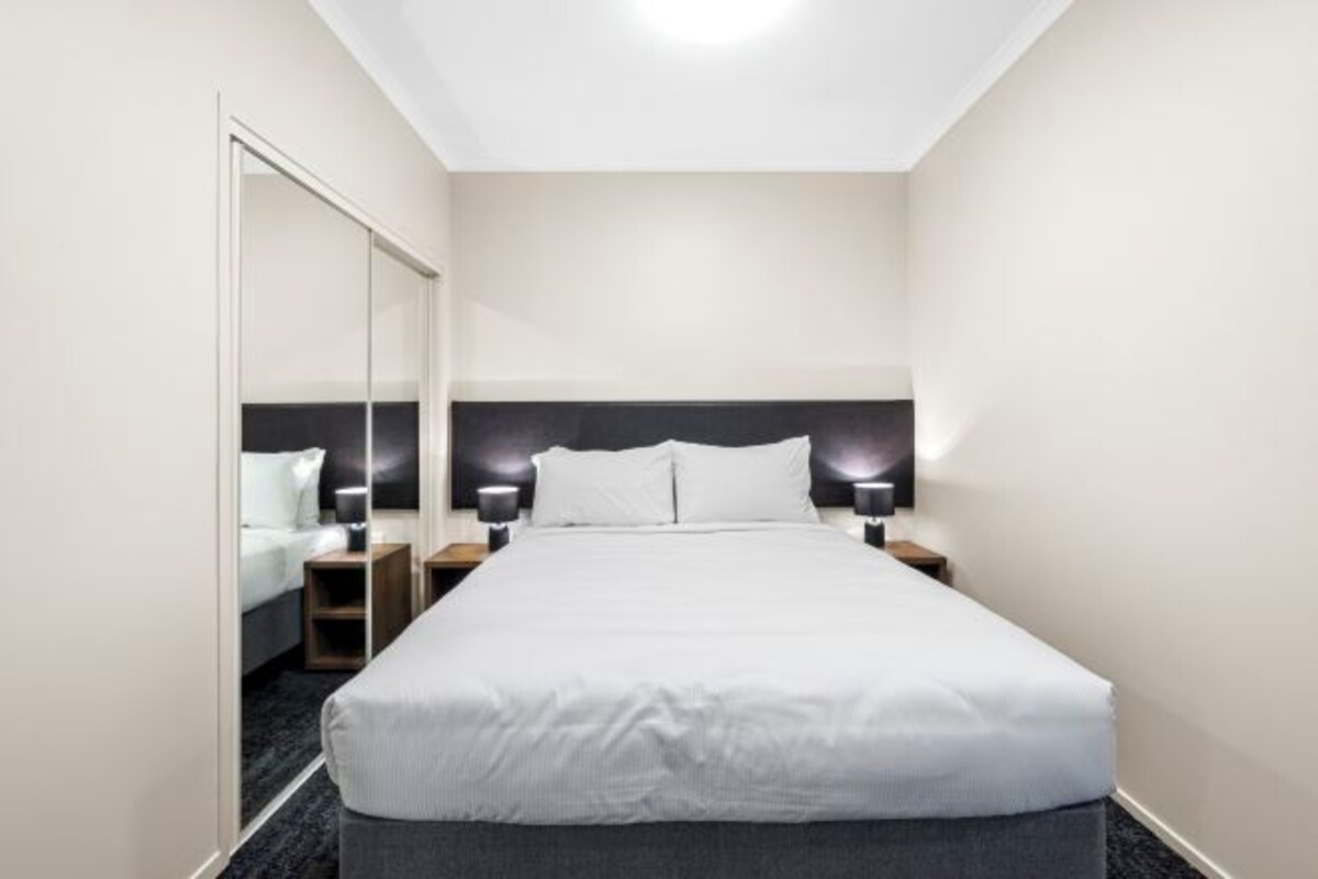 A hotel room with a large bed, two bedside tables with lamps, a mirror, and a dark headboard.