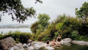 Una fuente termal natural con tres personas sentadas sobre rocas, rodeado de vegetación y vistas a un lago.
