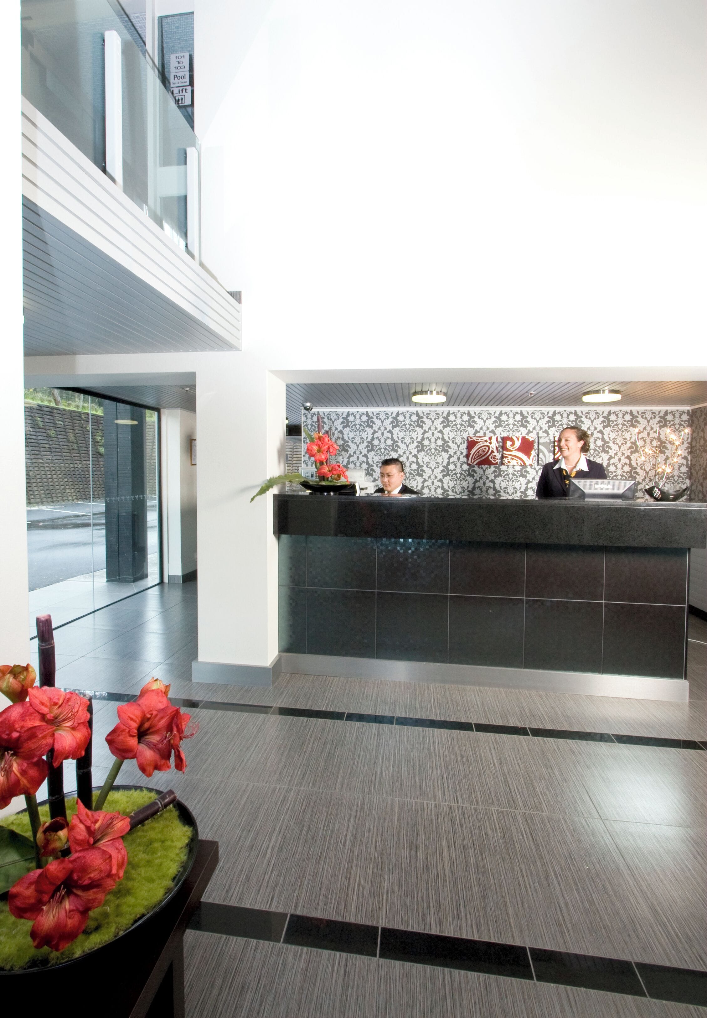 A modern reception area with a reception desk, two staff members, and a decorative plant with red flowers.