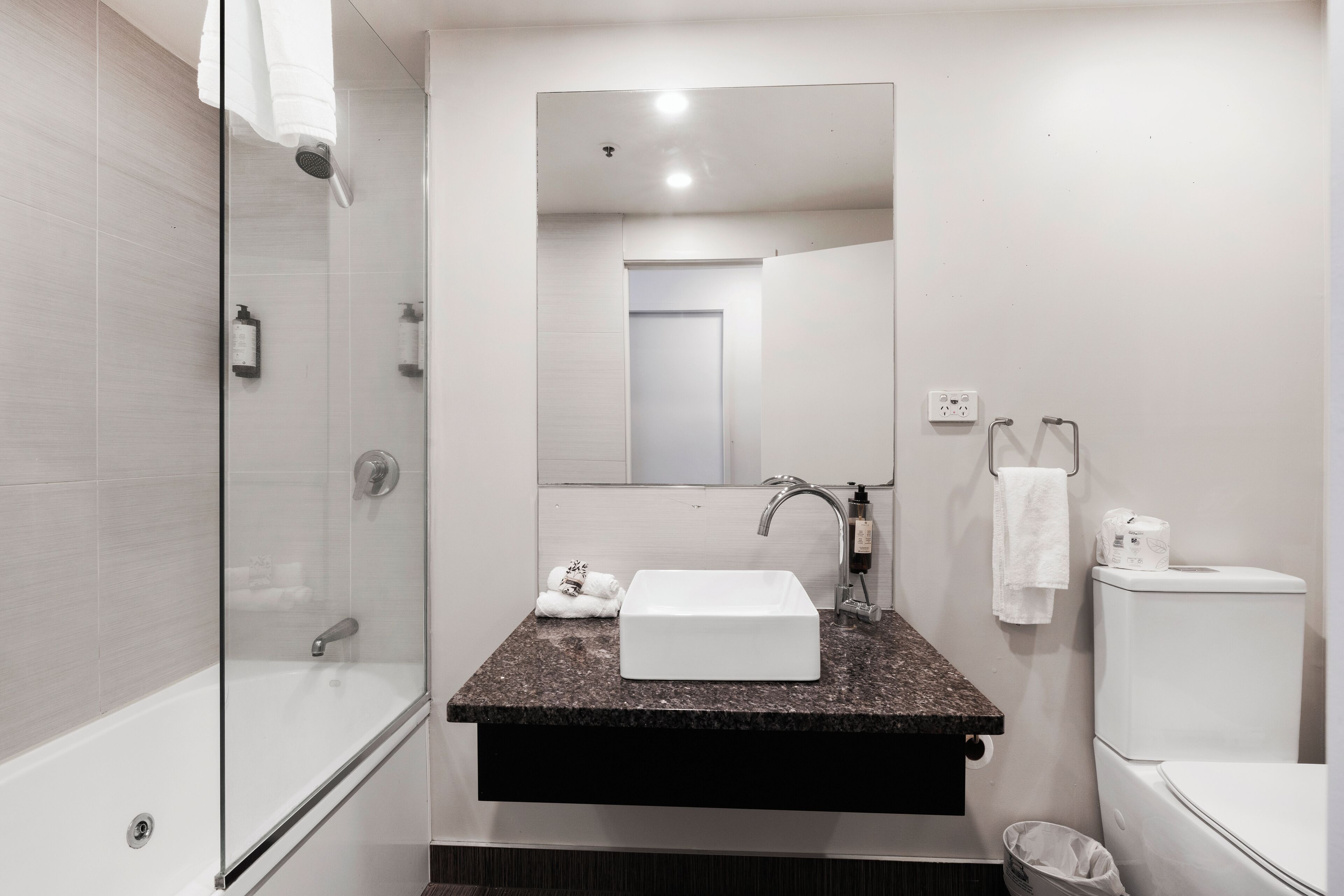A modern bathroom with a glass-enclosed shower, a white sink with a granite countertop, a mirror above the sink, and a toilet with a paper towel dispenser.