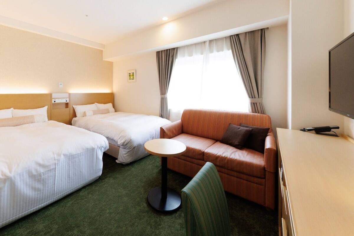 A hotel room with two beds, a sofa, a small table, a TV, and a window with curtains.