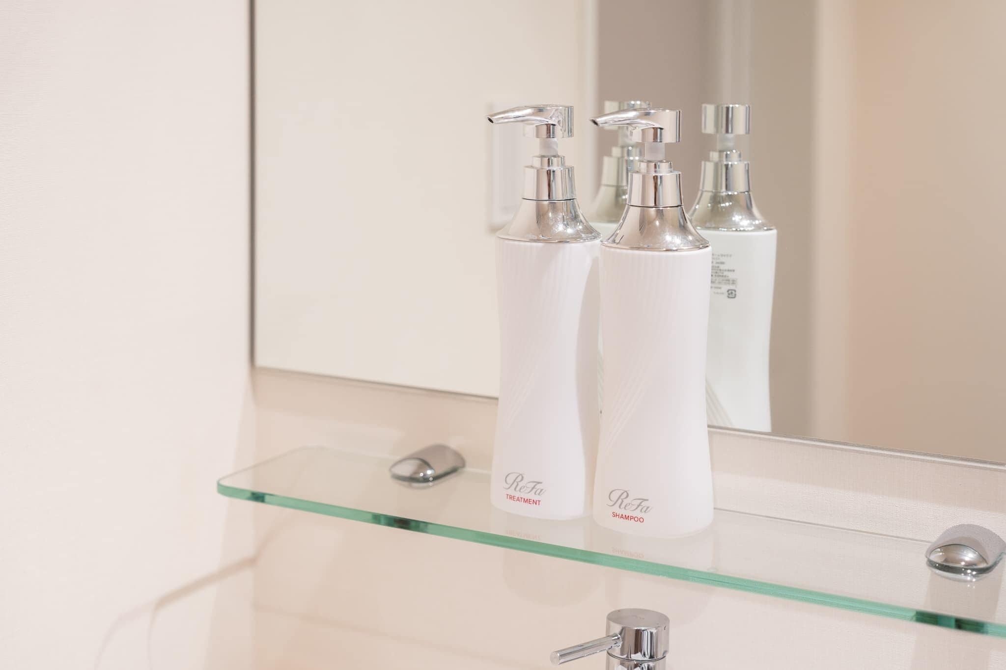 A bathroom with a glass shelf holding two white bottles labeled "ReFa Treatment" and "ReFa Shampoo" and a silver faucet.
