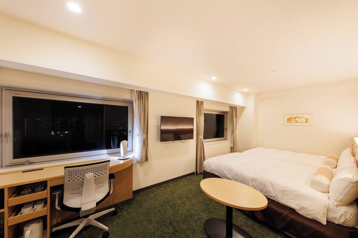 A hotel room with a large bed, a desk with a chair, a small table, a TV, and a window with curtains.