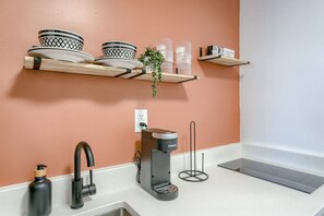 Una cocina con encimera blanca, una cafetera, un fregadero y una estantería en la pared con vajilla y una planta.