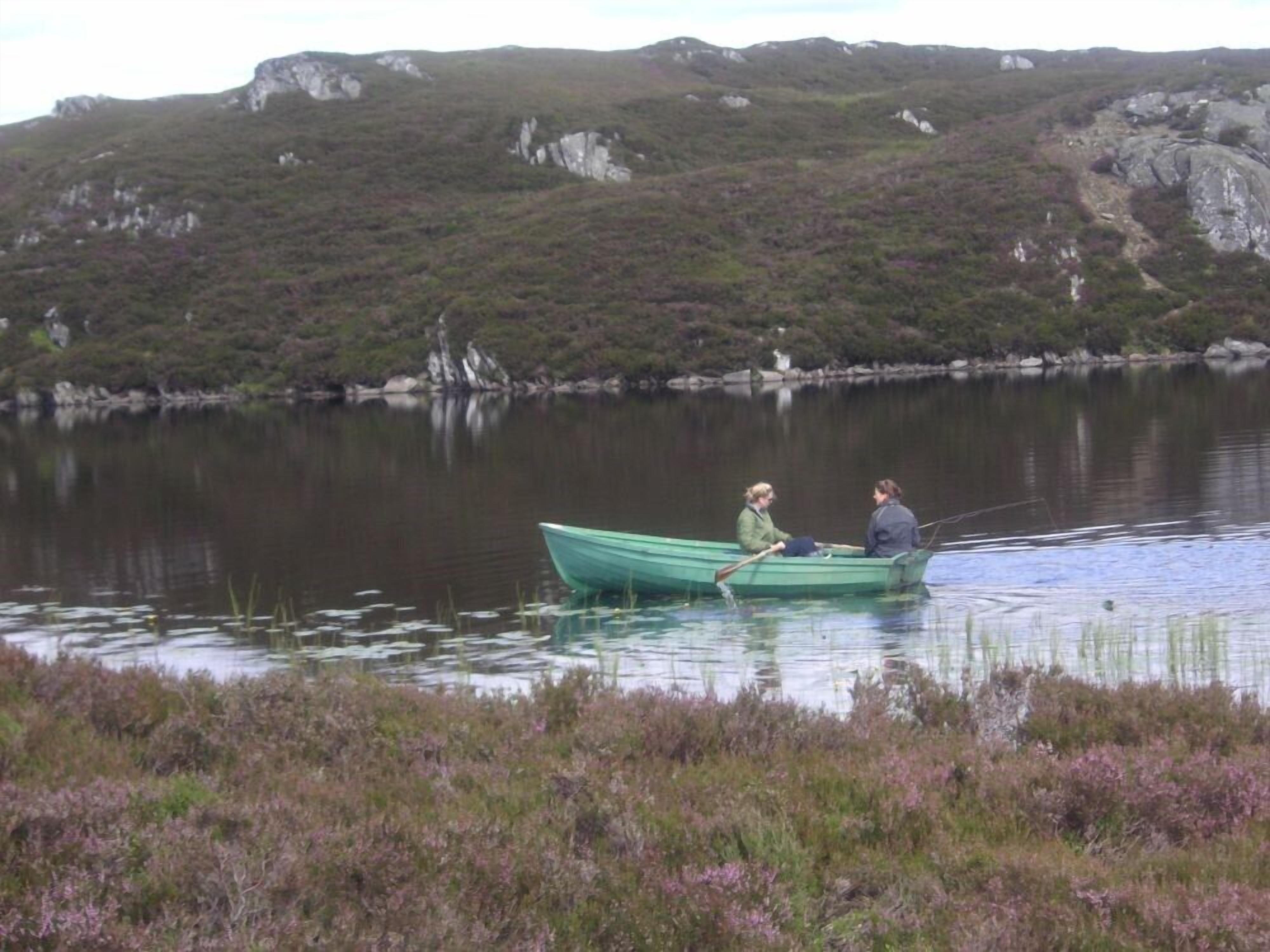 Two people in a small green boat on a calm lake surrounded by hills.