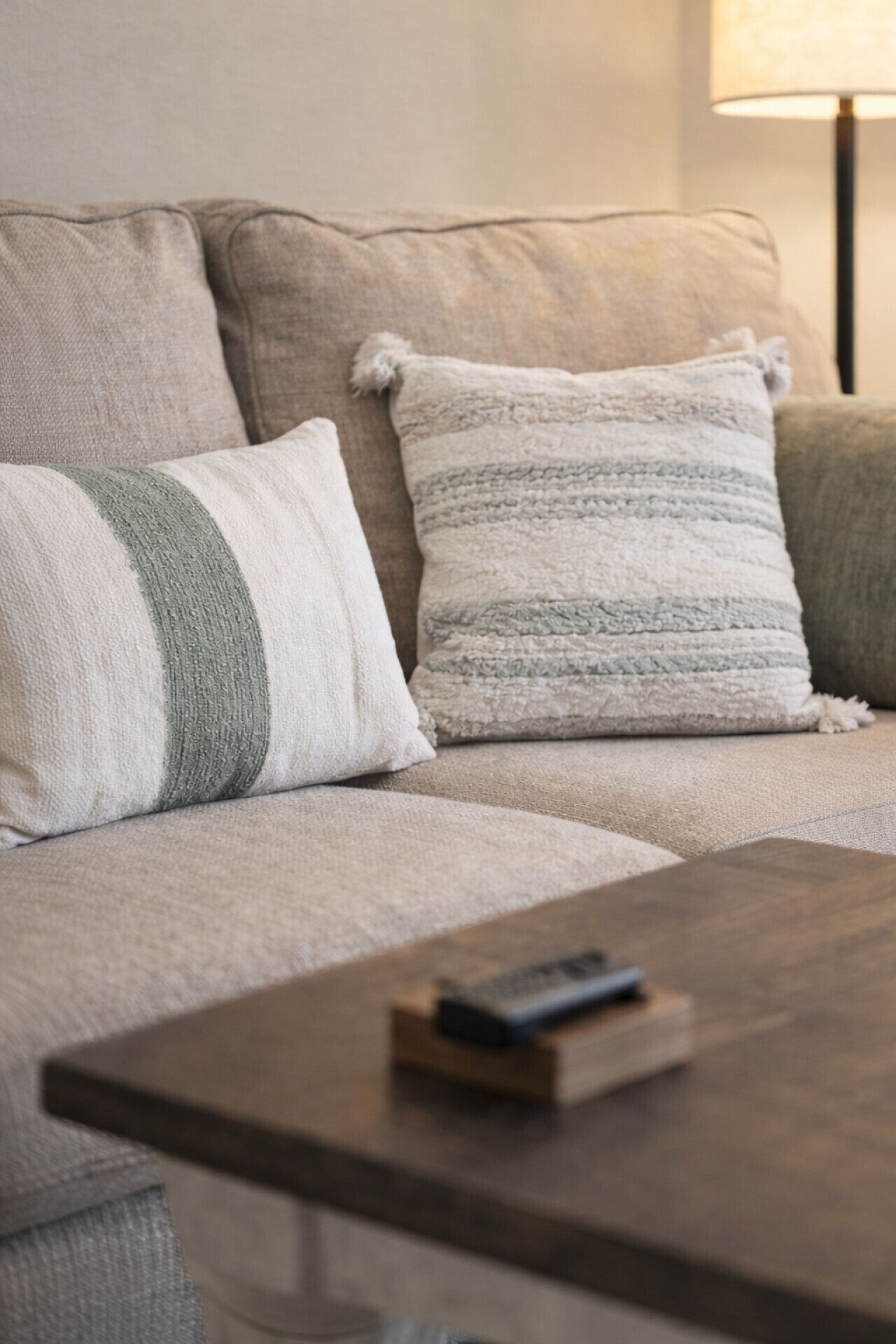 A beige sofa with two pillows, one striped and one solid, and a remote control on a wooden coffee table.