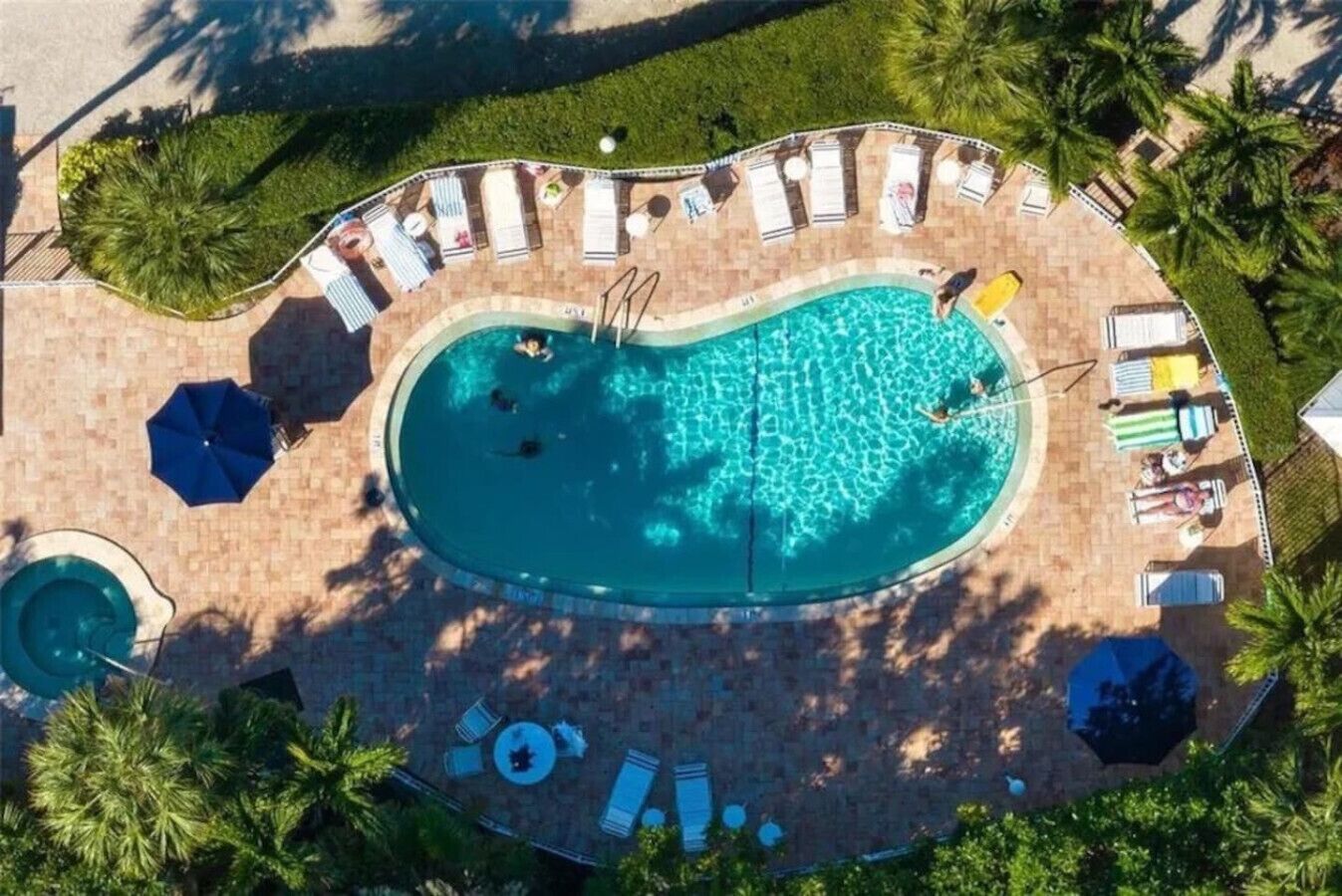 Une vue aérienne d’une piscine entourée de chaises longues et de parasols, avec des palmiers et une terrasse en briques.
