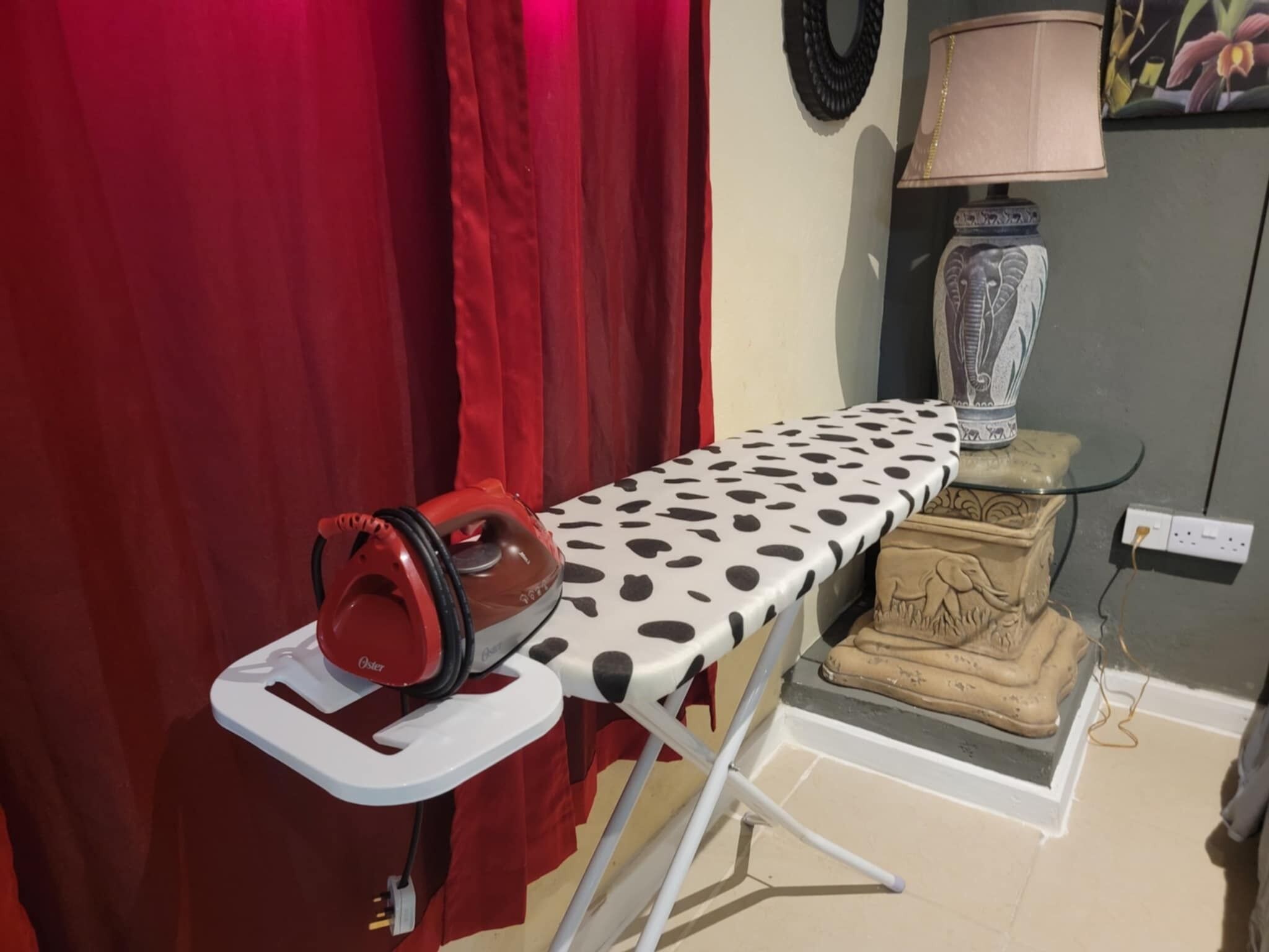 A room with a red curtain, an ironing board with a polka dot cloth, a red iron, a lamp, and a decorative statue.