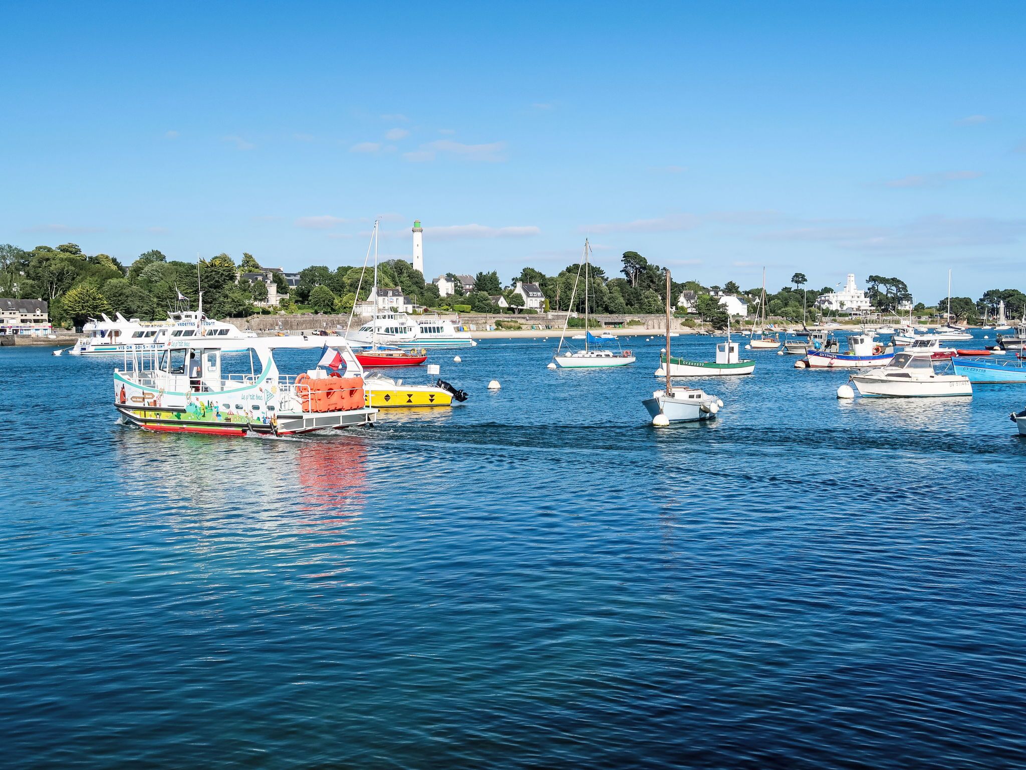 Un port de plaisance où sont amarrés différents types de bateaux, un phare en arrière-plan et une ville côtière avec des maisons et de la végétation.