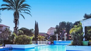 A swimming pool with clear blue water, surrounded by palm trees and other greenery, with buildings in the background. - Malibu Village by Interhome (Canet-Plage)