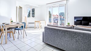 A modern living room with a grey sofa, a dining table with chairs, a TV, and a balcony with a view of buildings. - Malibu Village by Interhome (Canet-Plage)