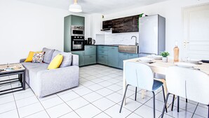 A compact living space with a kitchenette, a grey sofa with yellow cushions, a dining table with chairs, and a hanging light fixture. - Malibu Village by Interhome (Canet-Plage)