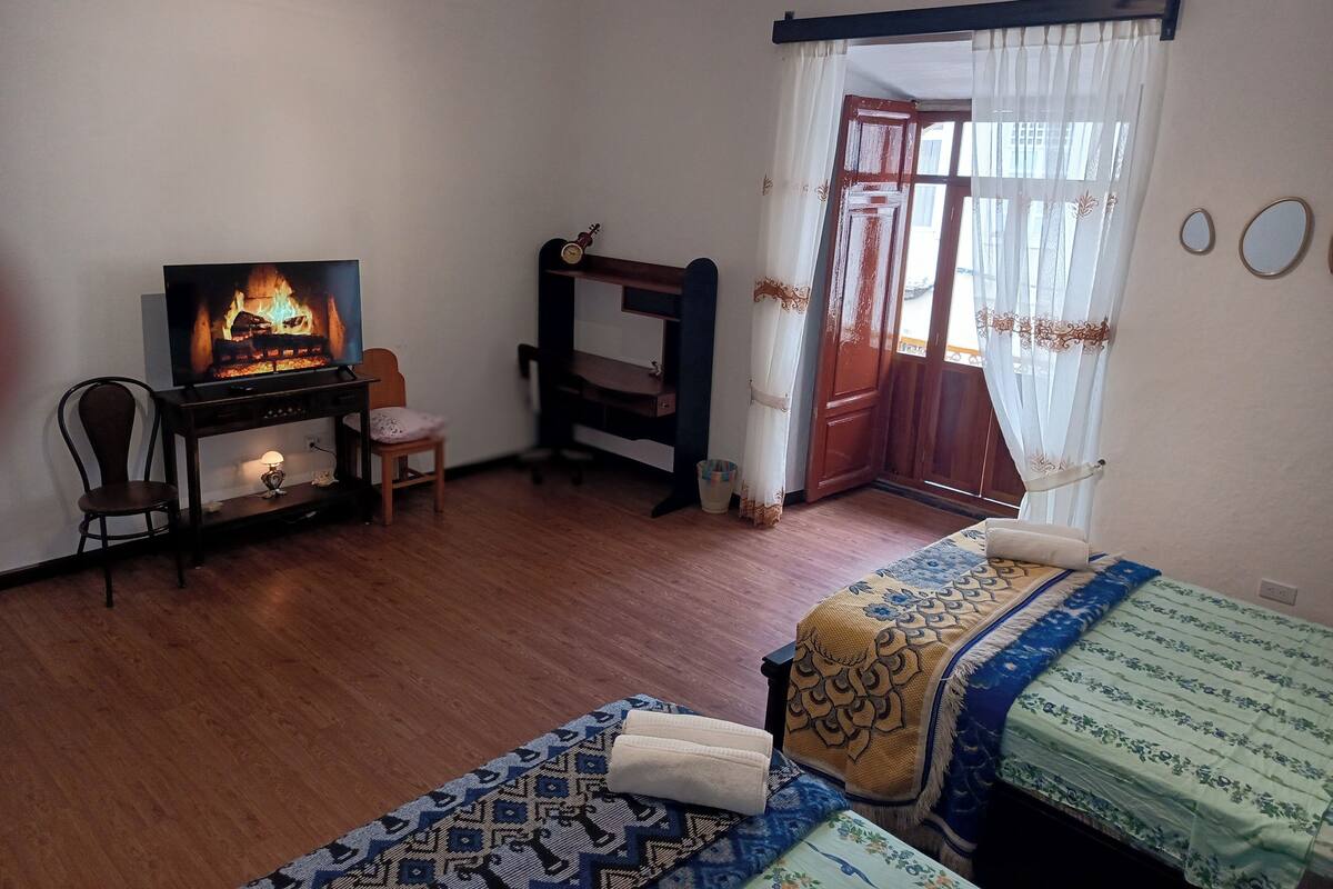 A room with a television displaying a fireplace, a desk with a chair, a bed with patterned bedding, and a window with curtains.